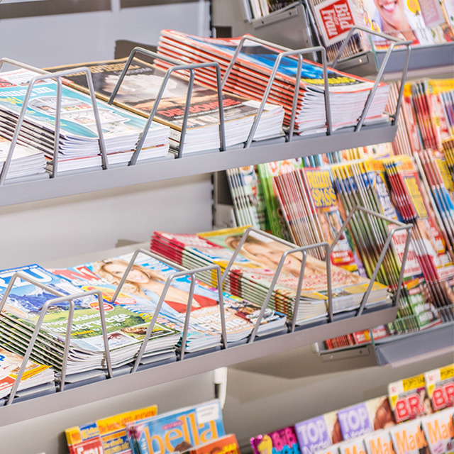 Ausschnitte eines beleuchteten Zeitungsregals mit vielen verschiedenen Zeitschriften, die in Stapeln nebeneinander liegen.