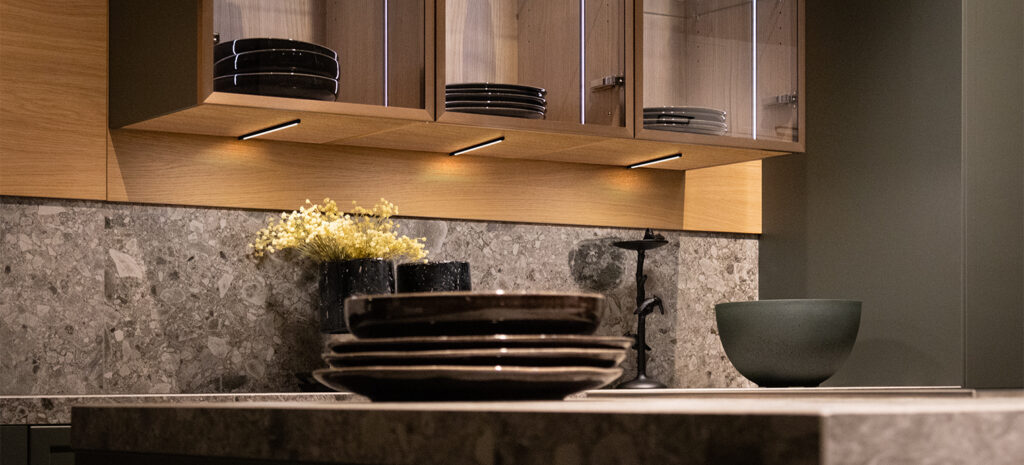 Küche mit grauem Spritzschutz und holzfarbiger Nischenverkleidung sowie beleuchteten Oberschränken. Im Vordergrund stehen Teller auf einer Kücheninsel