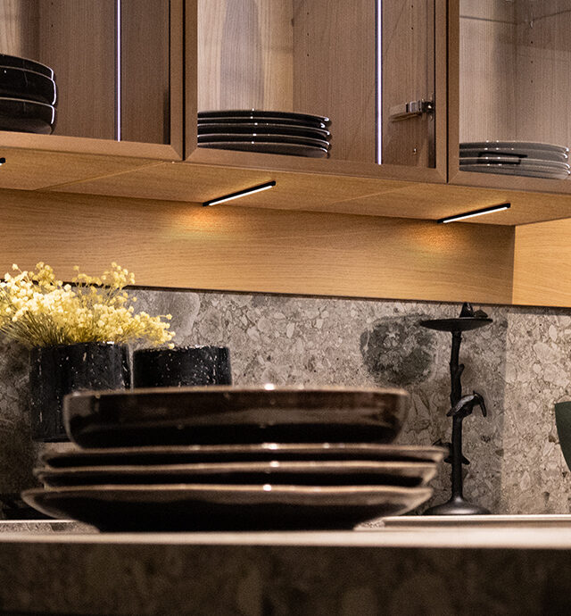 Küche mit grauem Spritzschutz und holzfarbiger Nischenverkleidung sowie beleuchteten Oberschränken. Im Vordergrund stehen Teller auf einer Kücheninsel