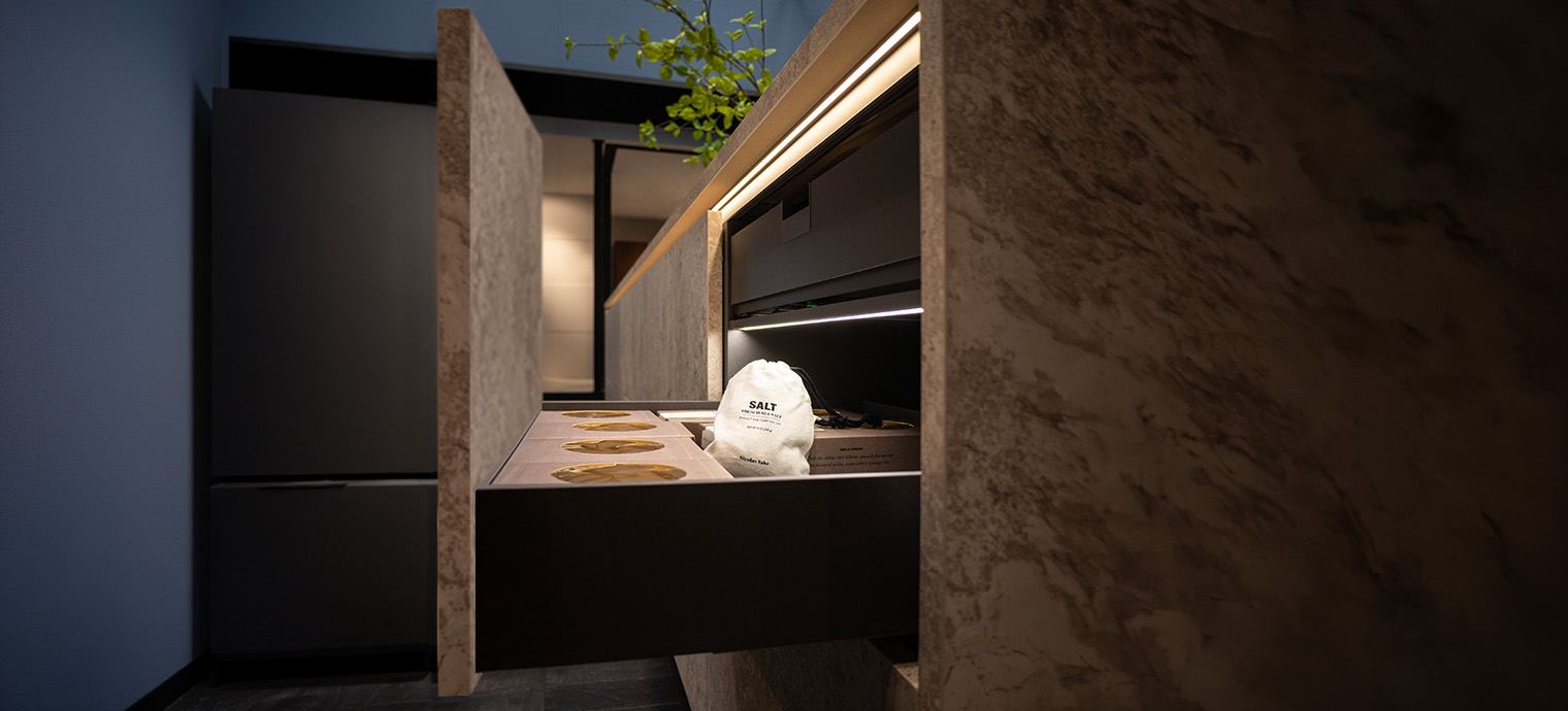 Eine moderne Kücheninsel mit einem offenen Schubladenfach, in dem ein Säckchen mit Salz und Verpackungen von Lebensmitteln von der LD 8132 Schubladenleuchte angestrahlt werden.