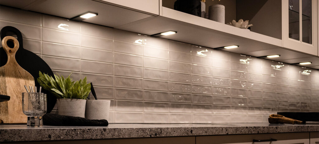 Eine moderne Küchenzeile mit weiß gefliester Rückwand und einigen Deko-Elementen auf der Arbeitsfläche. An den weißem Obschräknen wurden in regelmäßigen Abständen LD 8142 Leuchten angebracht, die sich stilvoll in das Ambiente integrieren und den Arbeitsbereich homogena ausleuchten.