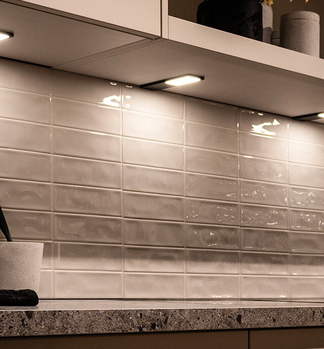 Eine moderne Küchenzeile mit weiß gefliester Rückwand und einigen Deko-Elementen auf der Arbeitsfläche. An den weißem Obschräknen wurden in regelmäßigen Abständen LD 8142 Leuchten angebracht, die sich stilvoll in das Ambiente integrieren und den Arbeitsbereich homogena ausleuchten.