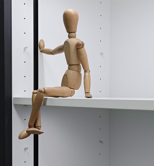 Eine Holz-Gleiderpuppe sitzt lässig auf einem weißen Regal, das einen modernen Raumakzent setzt.